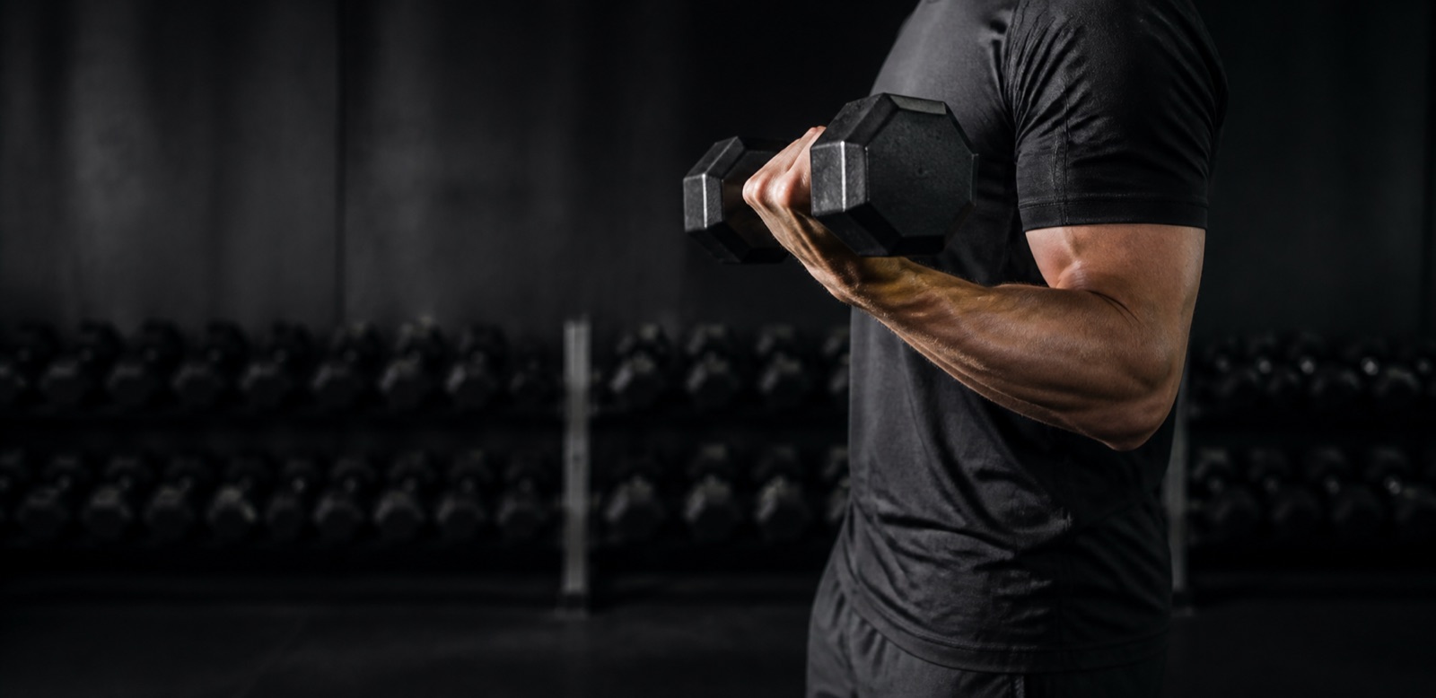 Person som utför hantelcurl i ett gym, sedd från sidan med hantlar i ett ställ i bakgrunden