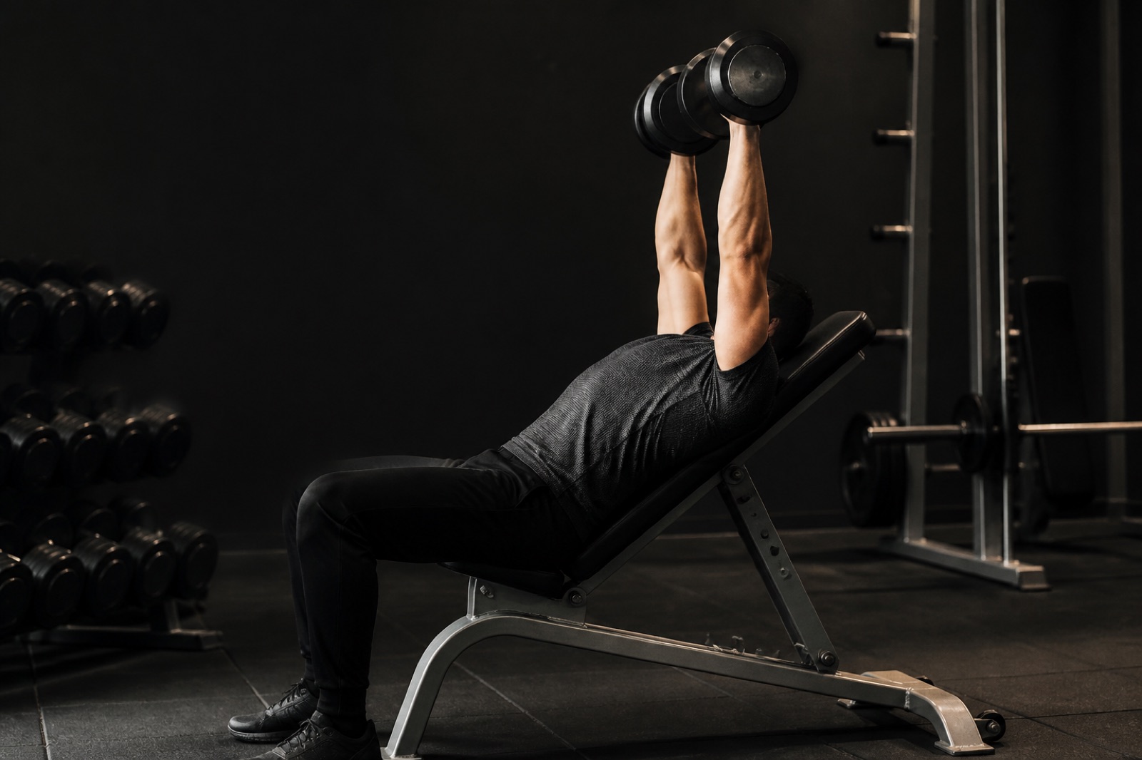 Person som utför lutande hantelpress på en bänk i ett gym, sedd från sidan