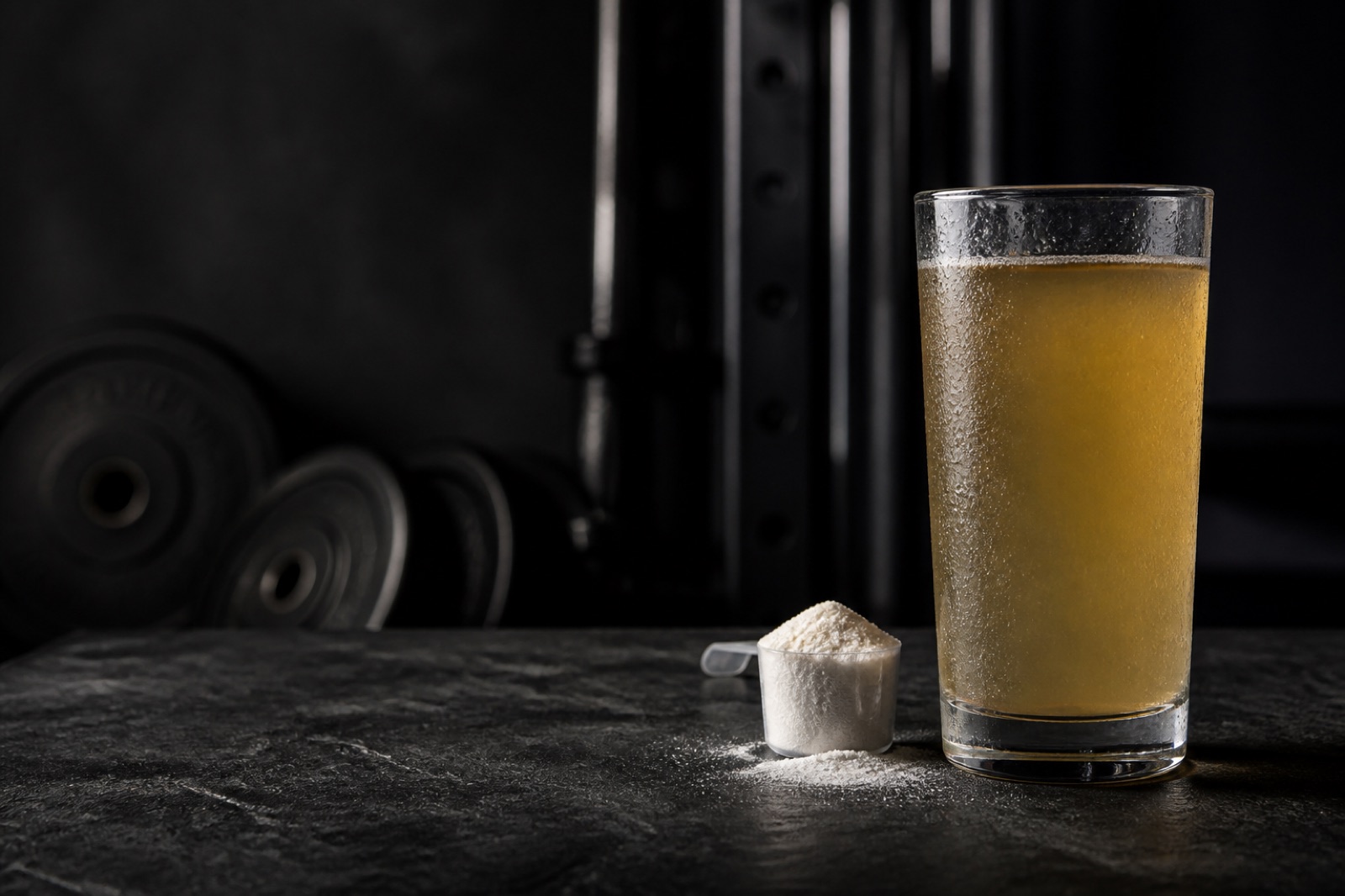 Högt glas med klar, saftliknande proteindryck på en mörk yta bredvid ett mått med pulver