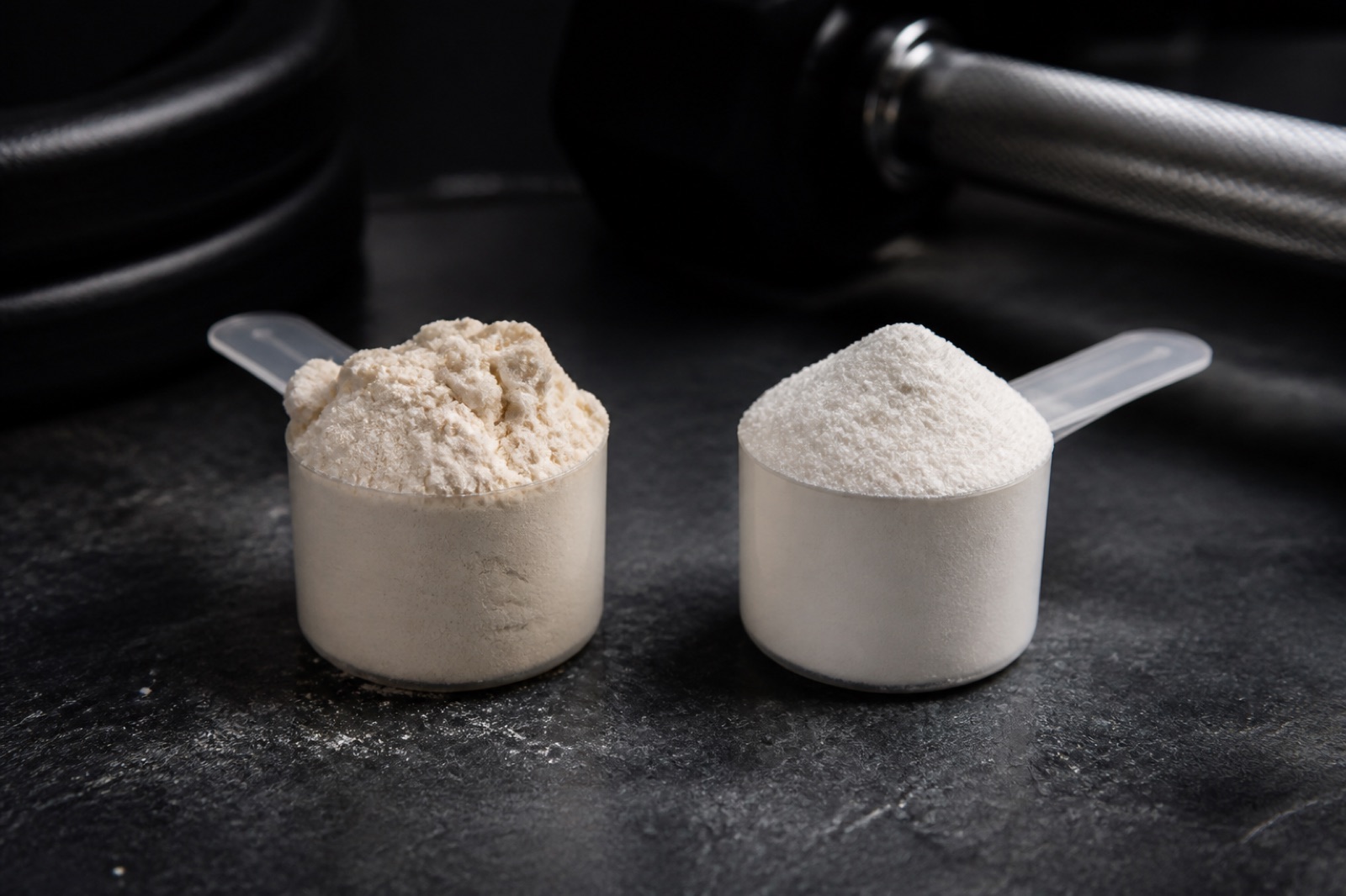 Två skopor vassleprotein sida vid sida på en mörk yta, ett grovkornigt koncentrat och ett finare isolat